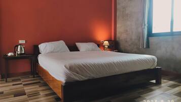 A neatly made bed in a room with a red wall, a window with curtains, a bedside table with a lamp, and a kettle on the floor.