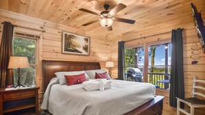 A wooden cabin bedroom with a bed, nightstands, a ceiling fan, and a window with curtains. - Mountain Views & just mins to City! Wood burning Fireplace, Hot tub, Fire pit  (Cherry Log)