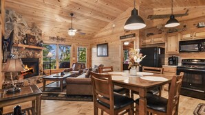 A cozy cabin interior with a fireplace, dining area, kitchen, and a living room with a sofa and chairs. - Mountain Views & just mins to City! Wood burning Fireplace, Hot tub, Fire pit  (Cherry Log)