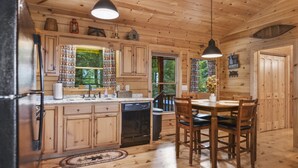 A wooden cabin interior with a kitchen and dining area, featuring wooden cabinets, a black refrigerator, a dining table with chairs, and hanging pendant lights. - Mountain Views & just mins to City! Wood burning Fireplace, Hot tub, Fire pit  (Cherry Log)