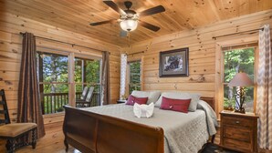 A wooden cabin bedroom with a bed, a ceiling fan, a nightstand, a lamp, and a window with curtains. - Mountain Views & just mins to City! Wood burning Fireplace, Hot tub, Fire pit  (Cherry Log)