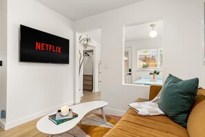 A modern living room with a flat-screen TV displaying 'NETFLIX,' a white coffee table with a book and candle, a brown sofa with a green pillow, and a window with blinds. - Luxe Studio Retreat - Close to DT & UT Austin (Austin)