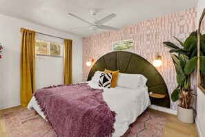 Une chambre à coucher avec un lit, un ventilateur de plafond, une fenêtre avec des rideaux, un luminaire fixé au mur et une plante en pot.