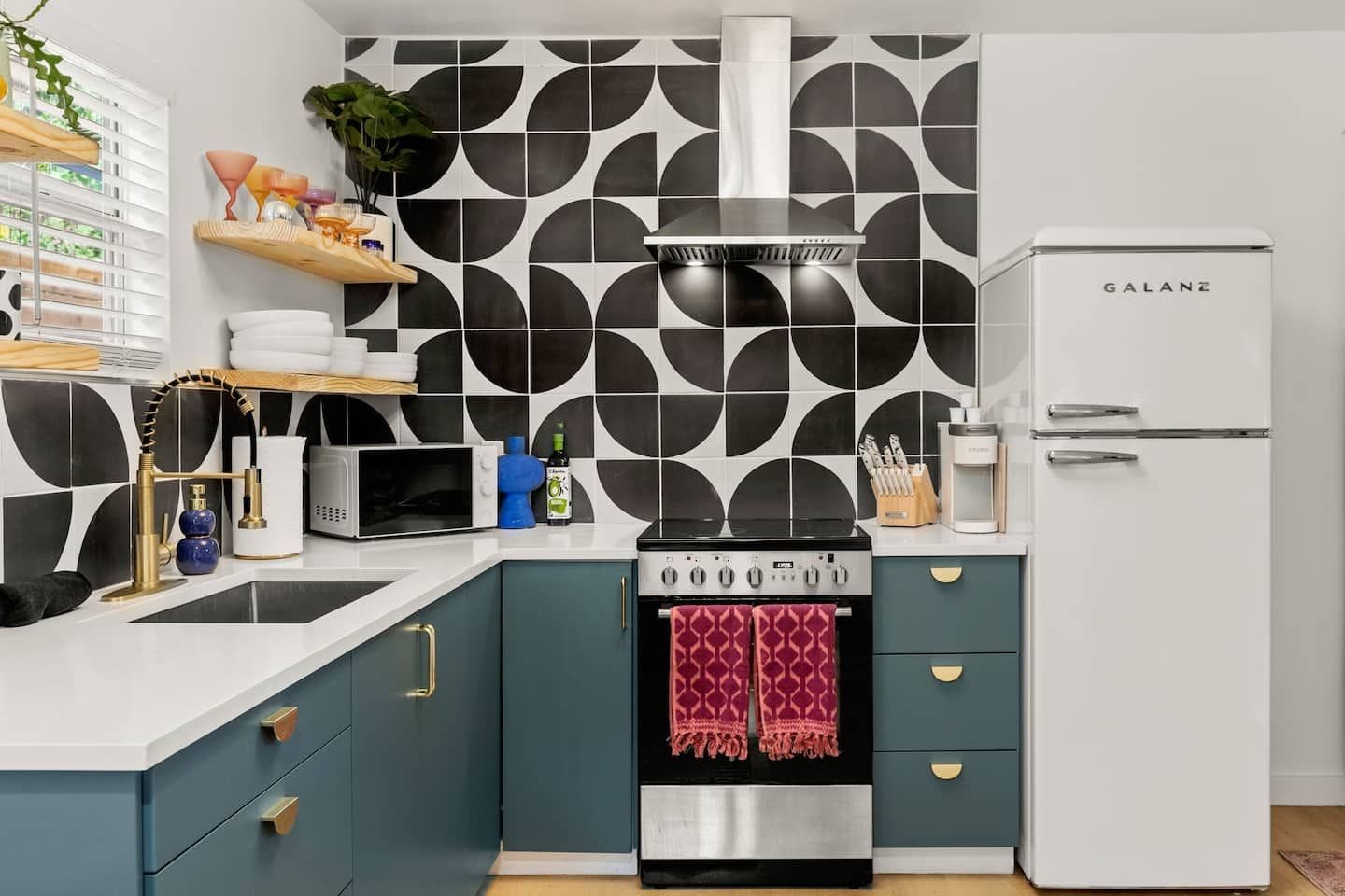 A modern kitchen with a black and white geometric backsplash, a stainless steel range hood, a white Galanz refrigerator, and teal cabinetry.