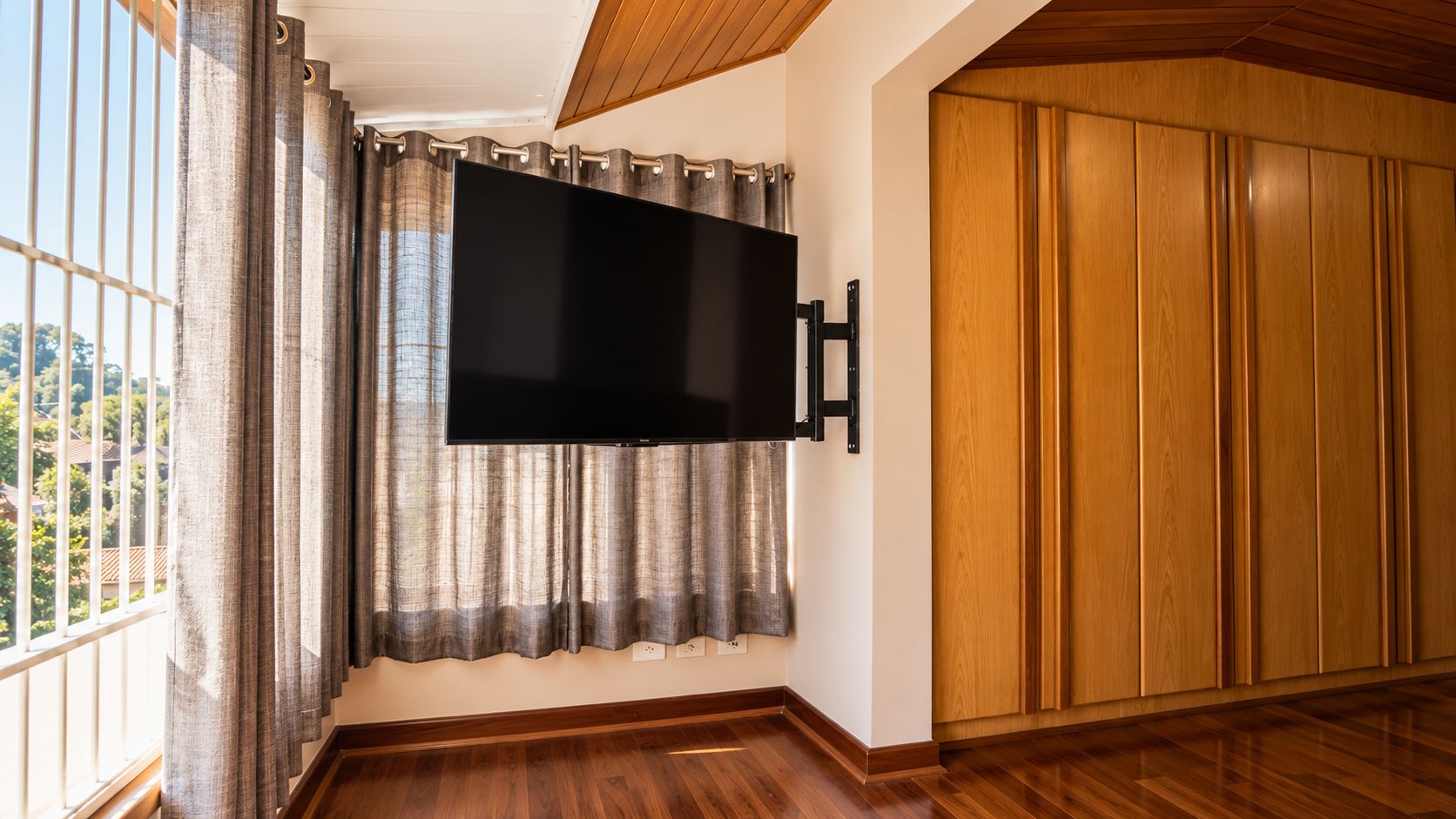 Ein Zimmer mit einem an der Wand befestigten Flachbildfernseher, Holzböden, einer großen Holztür und einem Fenster mit Vorhängen.