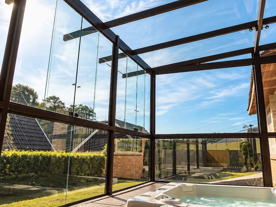 Área externa envidraçada com hidromassagem e espreguiçadeiras, com vista para um jardim e um edifício.