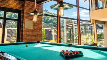 A pool table with balls, hanging pendant lights, and a view of the outdoors through large windows.