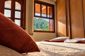 A bedroom with a large window, a bed with a red pillow, and a view of a forest. - Pousada Atalaia Village (Campos do Jordão)