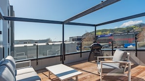 A balcony with a view of a cityscape, including a train station and buildings. There is a grill and a table with chairs. - Inner City Apartment 2 with Car Park + Rooftop Balcony (Nelson)