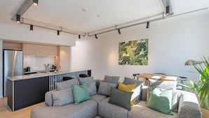 A modern living room with a grey sectional sofa, a small black coffee table, and a kitchen area with a stainless steel refrigerator and a bar counter. - Inner City Apartment 2 with Car Park + Rooftop Balcony (Nelson)