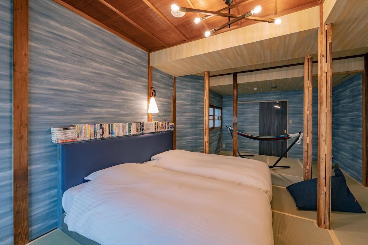 A modern bedroom with a large bed, a bookshelf, a hammock, and a view of a room through a glass wall.