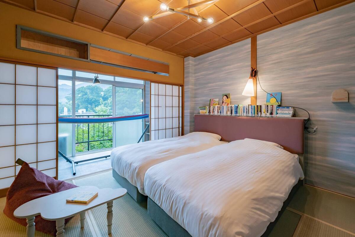 A hotel room with a large bed, a small table, a chair, a bookshelf, and a view of greenery outside.