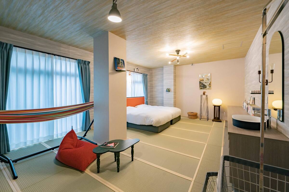A hotel room with a bed, a hammock, a small table, a red bean bag, a bedside table, a mirror, a sink, and a ceiling fan.