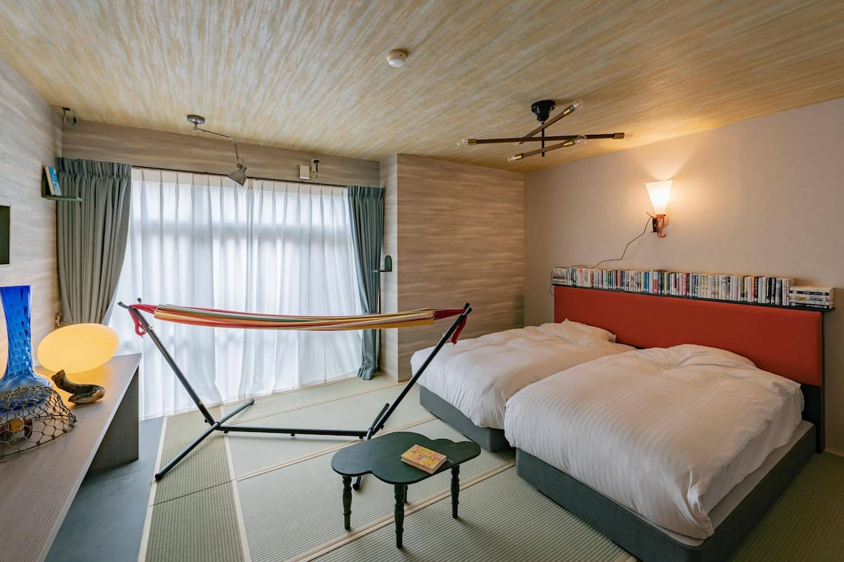 A modern hotel room with a large bed, a small table, a hammock, and a bookshelf.