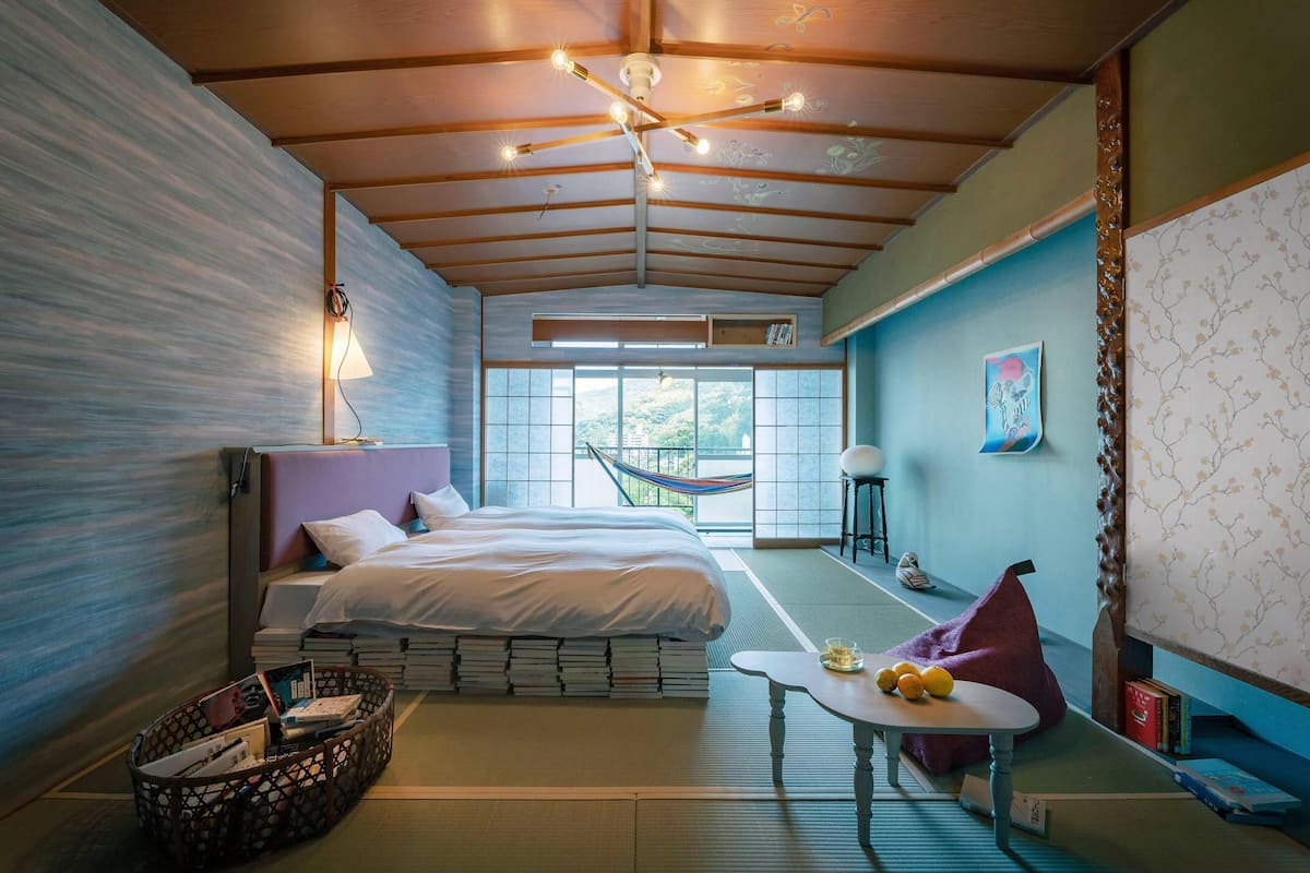 A modern hotel room with a large bed, a small table, a hammock, and a view of the outdoors.
