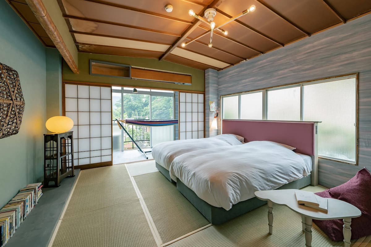 A bedroom with a bed, a small table, a chair, a bookshelf, and a view of the outdoors.