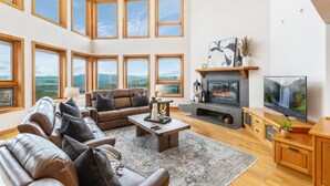 Ein geräumiges Wohnzimmer mit Kamin, Ledersofas, einem Fernseher und großen Fenstern mit Blick ins Freie.