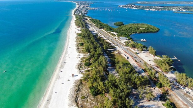 Une vue aérienne d’une zone côtière, avec une longue plage de sable, une route qui longe le rivage et une petite île couverte d’une végétation luxuriante.