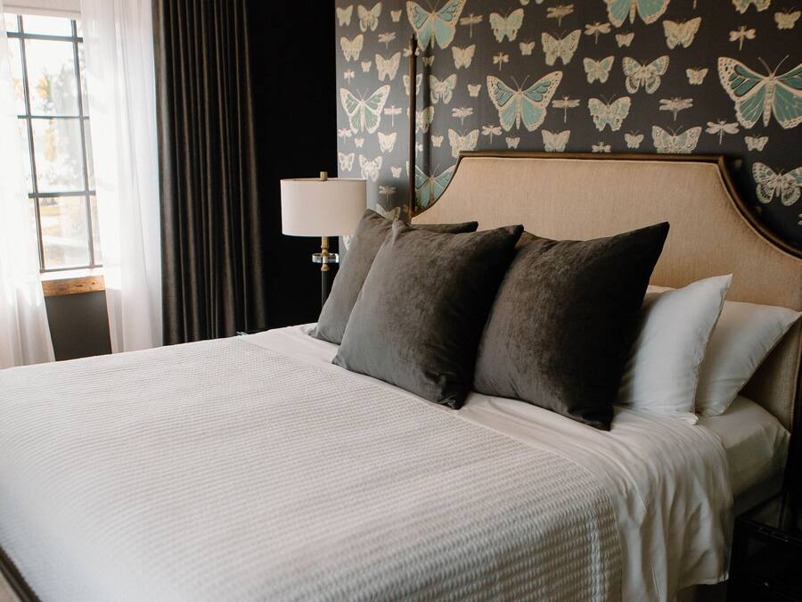 Cama bem arrumada com cabeceira, uma janela grande com cortinas e uma parede com padrão de borboletas.