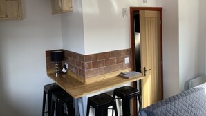 Une cuisine compacte dotée d’un plan de travail en bois, de deux tabourets de bar noirs, d’une petite étagère et d’une porte donnant sur une autre pièce.