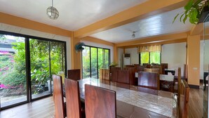 Un comedor espacioso con una gran mesa y numerosas sillas de madera, ventanas amplias que ofrecen vistas a la vegetación, y un ventilador de techo.