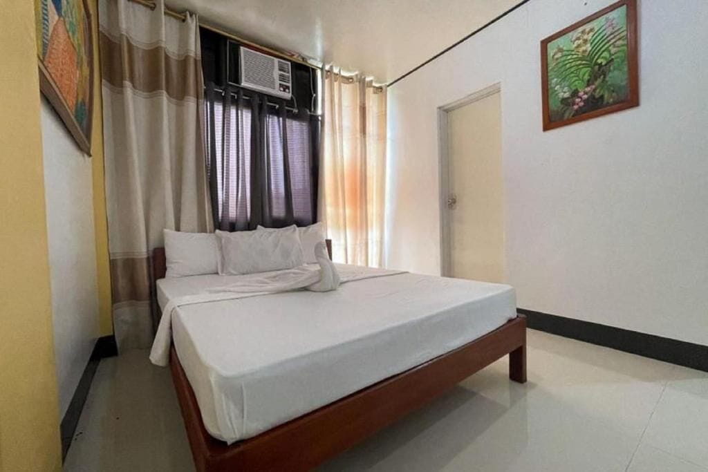 A single-bed room with a wooden frame, white bedding, a window with curtains, an air conditioning unit, and a door.