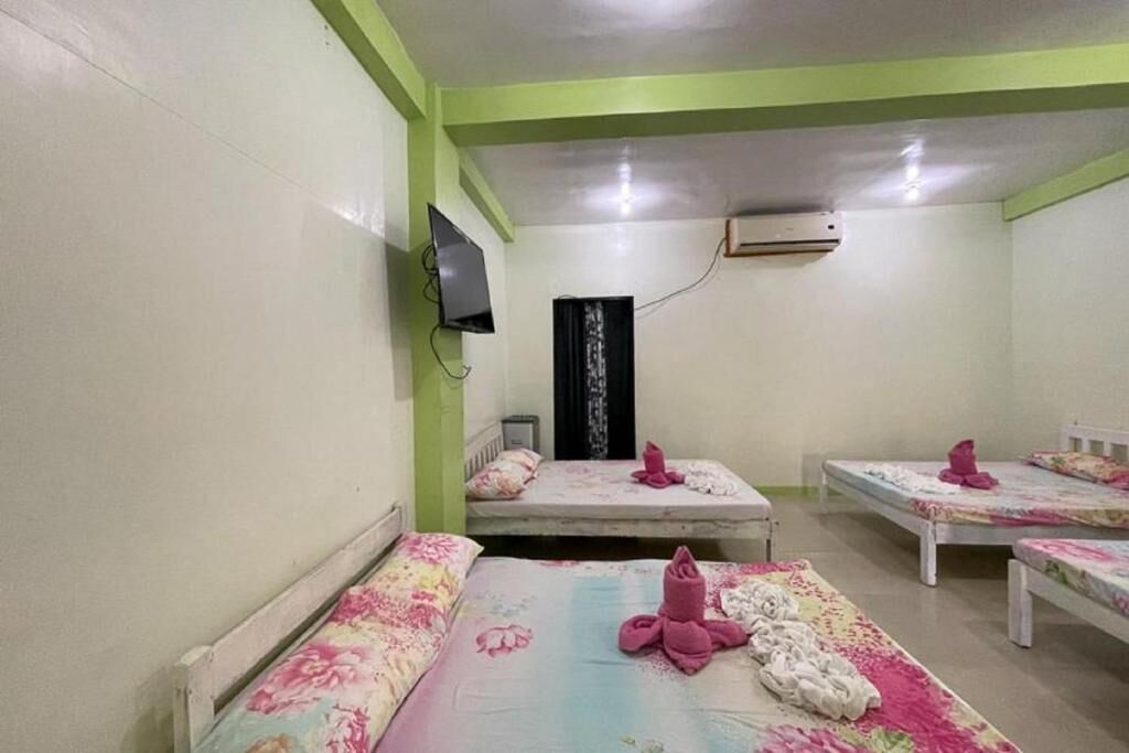 A room with four beds, each with floral bedding and pink towels. There is a television mounted on the wall and an air conditioning unit above it.