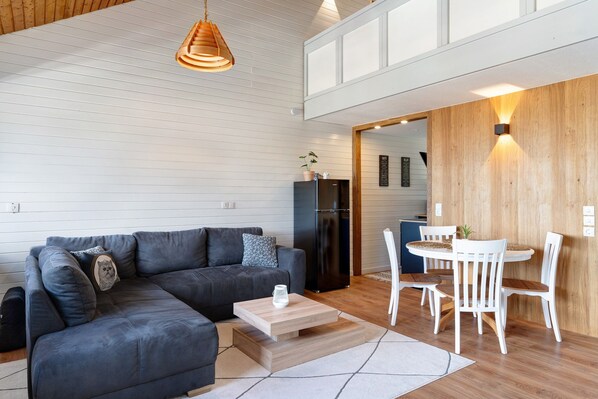 A modern living room with a sectional sofa, a dining area with white chairs, a wooden accent wall, and a kitchen in the background.