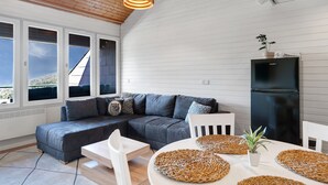 Un espace de vie moderne comprenant un canapé d’angle, une table à manger avec des sets de table en fibres naturelles, un réfrigérateur noir et de grandes fenêtres offrant une vue dégagée.