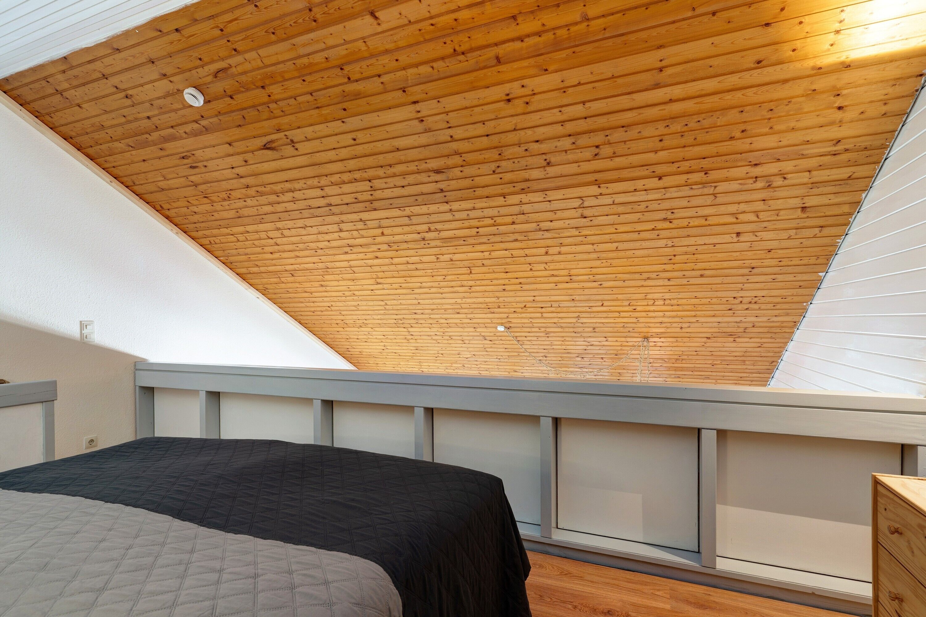 Ein Schlafzimmer mit schrägem Dach, einem Bett, einer Kommode und einem Fenster.