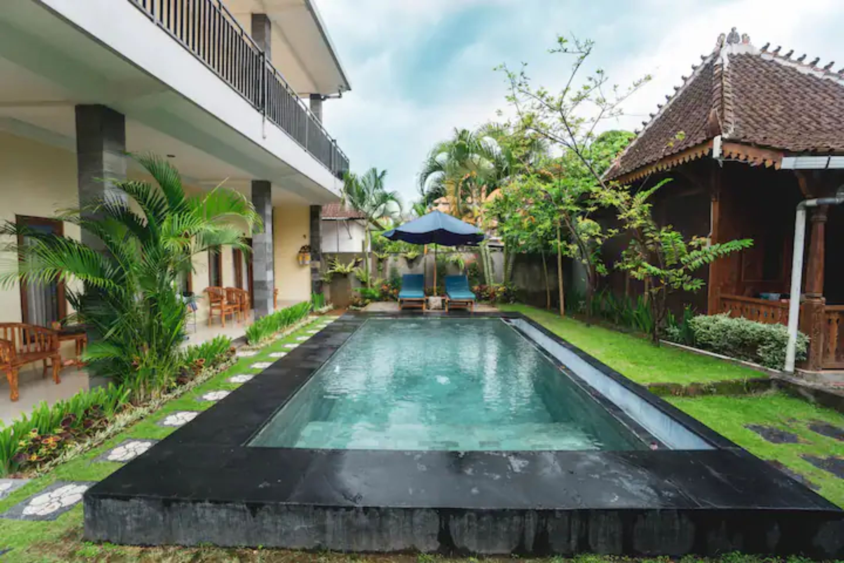 A swimming pool with a black border, surrounded by a garden with palm trees and a wooden structure with a tiled roof.