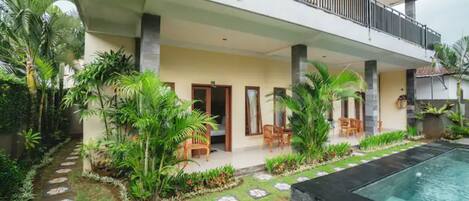 A two-story house with a balcony, a swimming pool, and a garden with tropical plants.