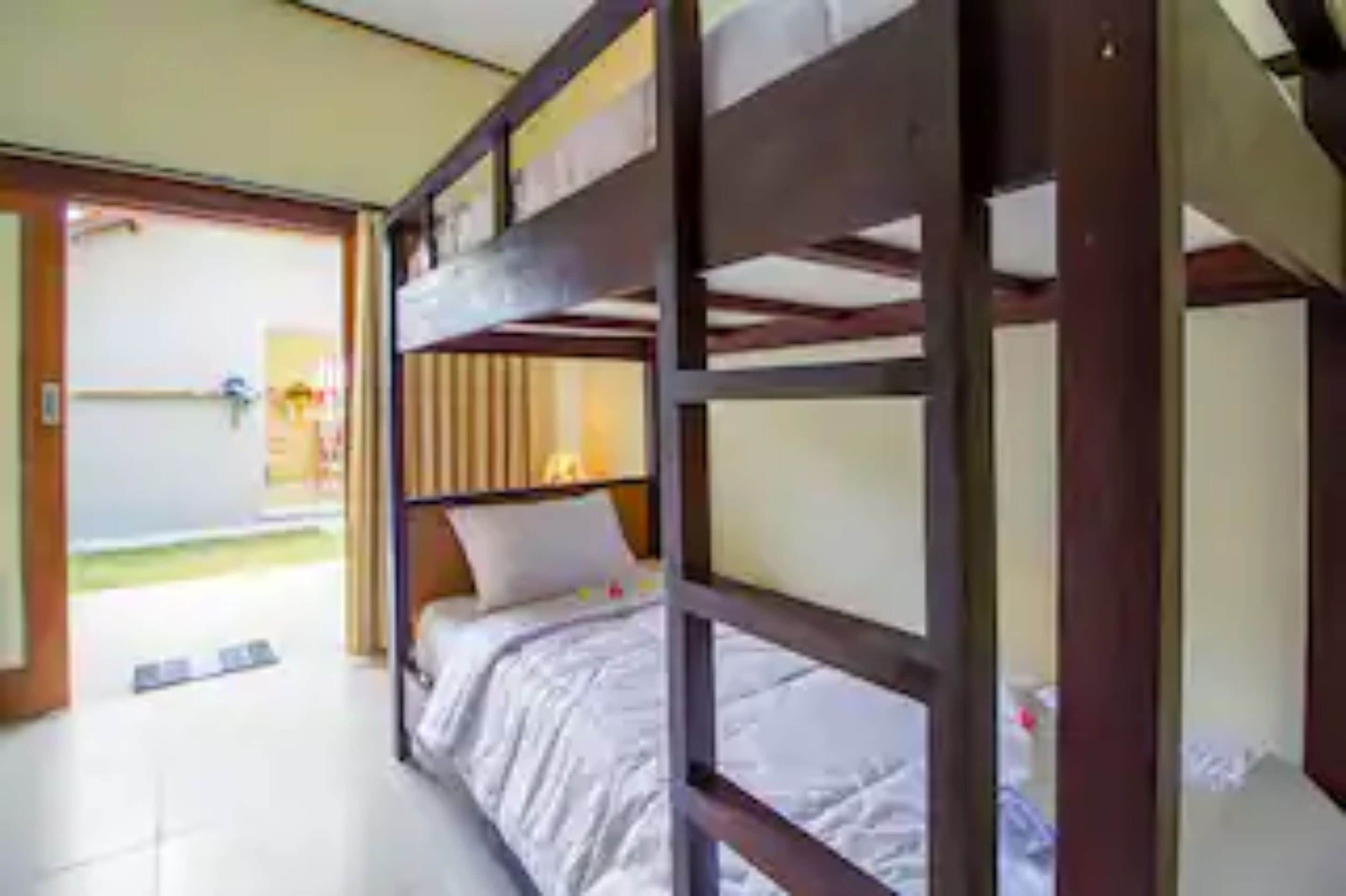 Une chambre avec un lit superposé, une structure en bois, un matelas blanc et un oreiller. Une porte donne accès à un balcon.