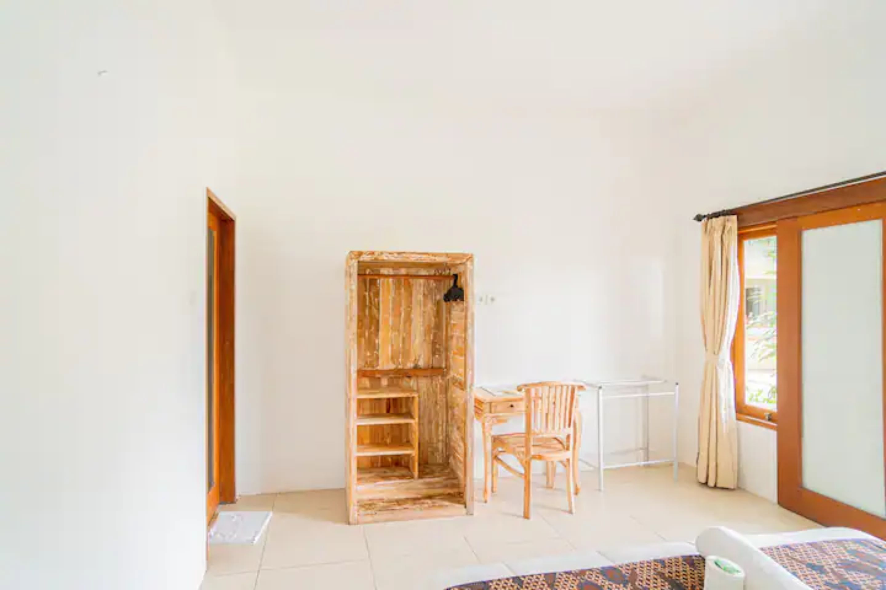 A room with a wooden wardrobe, a chair, a small table, and a bed.