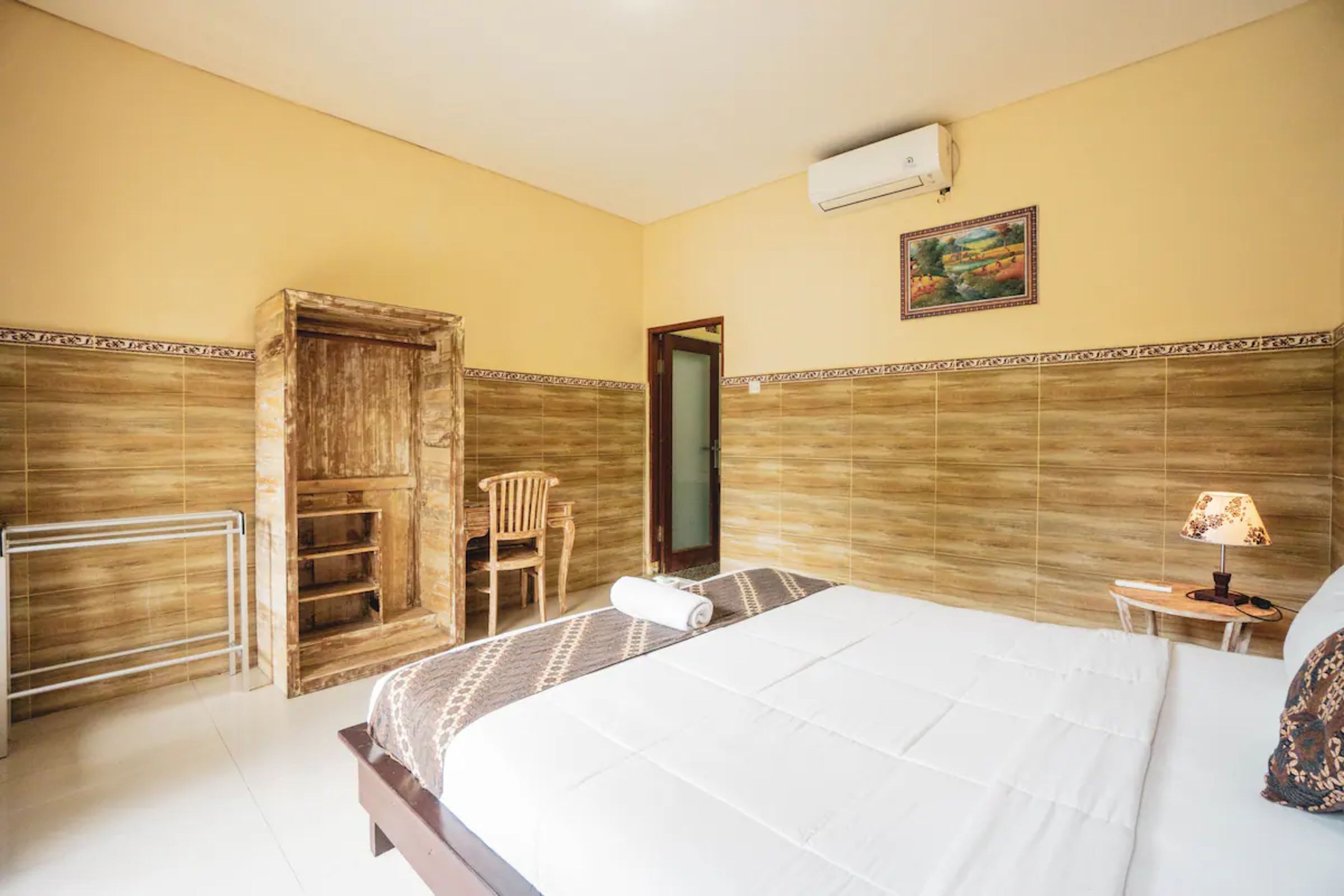 A hotel room with a bed, a wooden wardrobe, a chair, a small table with a lamp, and a painting on the wall.