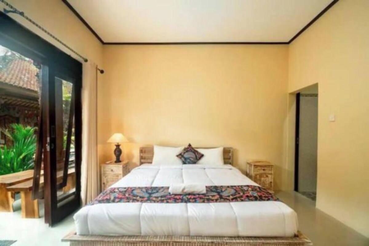 A bedroom with a bed, a nightstand, a lamp, and a view of the outside.