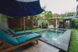 A swimming pool with lounge chairs and an umbrella, surrounded by greenery and a house in the background. - Pondok Lulik Homestay Canggu (Canggu)