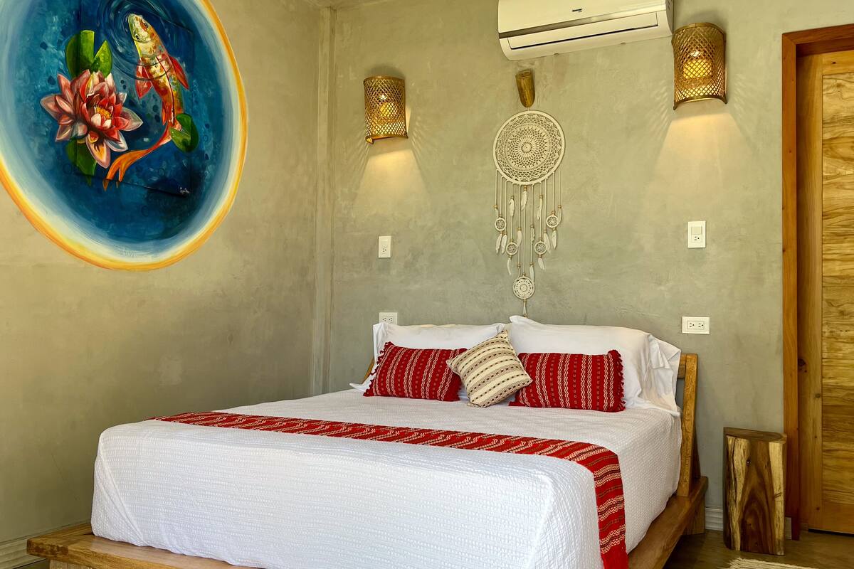 A bedroom with a bed, a wall-mounted air conditioner, two wall sconces, a dreamcatcher, and a painting of a fish and lotus.