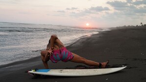 Una persona recostada sobre una tabla de surf en la playa, con el sol poniéndose en el horizonte.