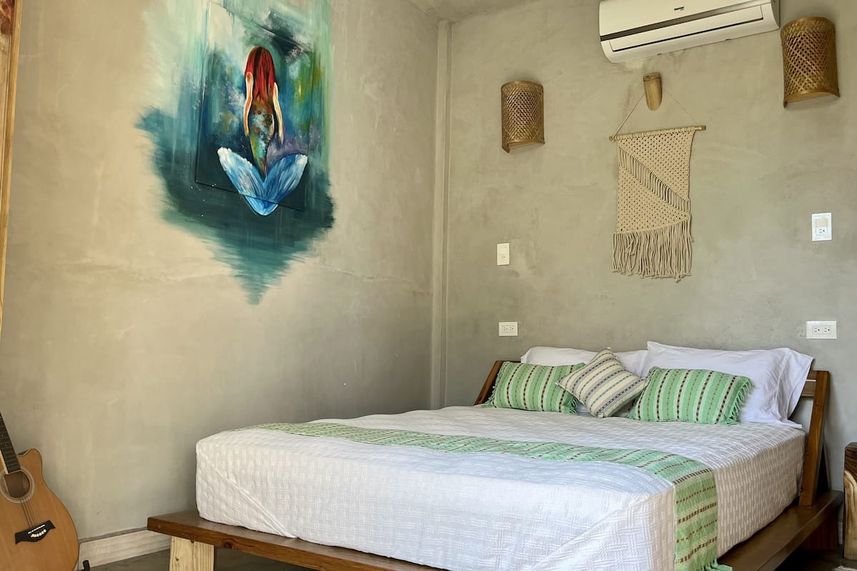 A bedroom with a bed, a guitar, a wall painting, and a macrame wall hanging.