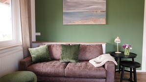 A living room with a brown sofa, green cushions, a painting, a small table with a lamp, and a window with curtains. - Apartment Säntis, 45 sqm with Balcony and Wi-Fi (Lindau am Bodensee)
