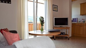 Een woonkamer met een bank, een rond bijzettafeltje, een televisie, en een keukenhoek met kastjes en een raam.