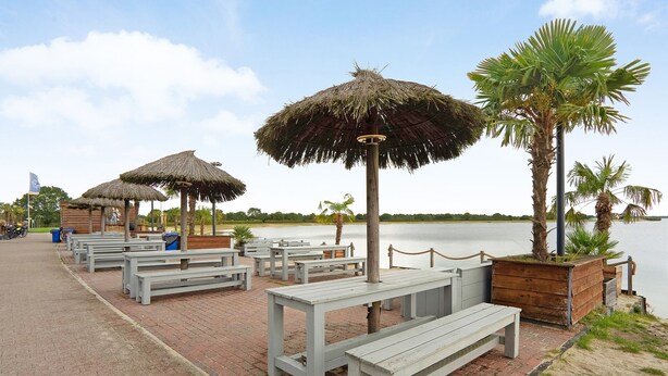Eine Seeterrasse mit Reetdachschirmen, Holzbänken und Blick auf den See.