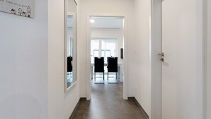 Ein schmaler Flur mit einer Tür rechts, einem Spiegel links und einem Essbereich mit Stühlen und Tischen am Ende.