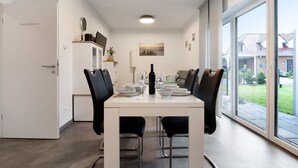 Ein Essbereich mit Tisch für vier Personen, Stühlen, einem weißen Schrank mit einer Uhr und einem großen Fenster mit Blick auf eine Wohngegend.