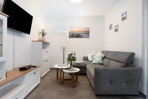 Un espace de vie compact comprenant un canapé gris, un téléviseur à écran plat fixé au mur, une petite table ronde avec un vase et un meuble orné d’une plante.