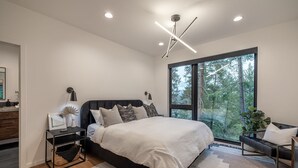 A modern bedroom with a large bed, bedside tables, a nightstand, a window with a forest view, and a ceiling light fixture.