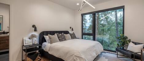 Une chambre moderne avec un grand lit, des tables de chevet, une commode, une fenêtre donnant sur une forêt et un luminaire au plafond.