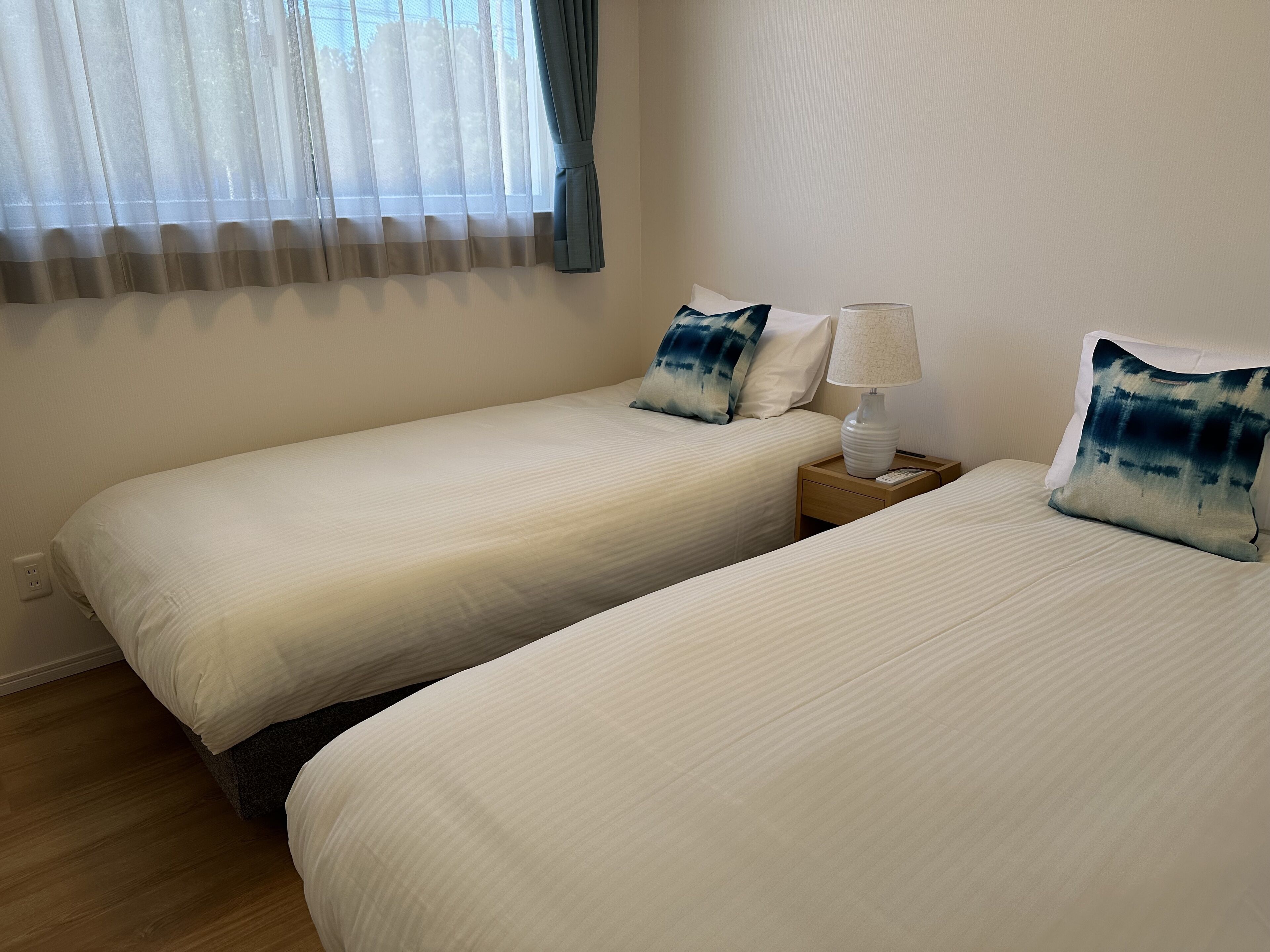 A hotel room with two single beds, a nightstand with a lamp, and a window with sheer curtains.