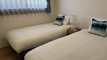 A hotel room with two single beds, a nightstand with a lamp, and a window with sheer curtains.